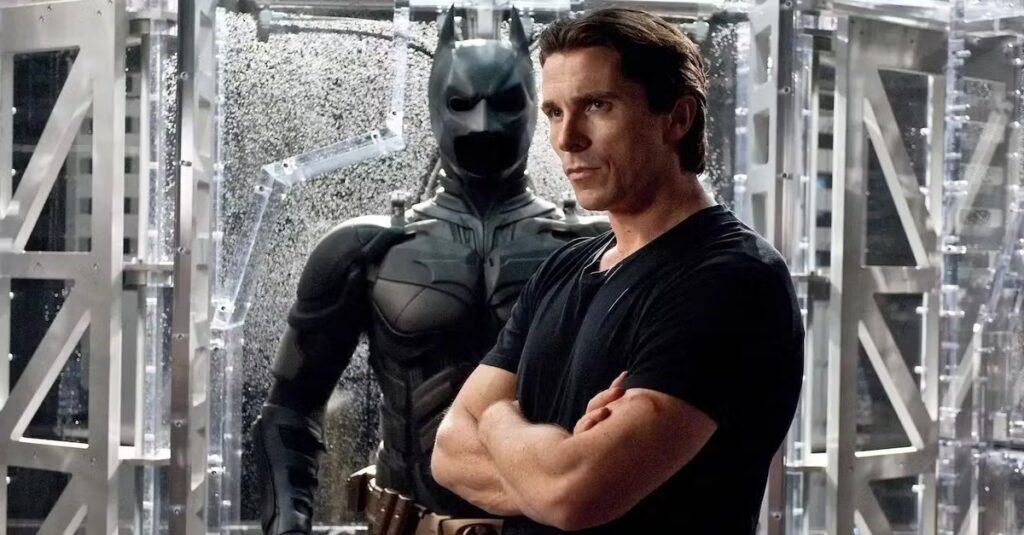 Scène du film Batman Begins. Bruce Wayne est debout les bras croisés devant un prototype de la tenue de Batman.